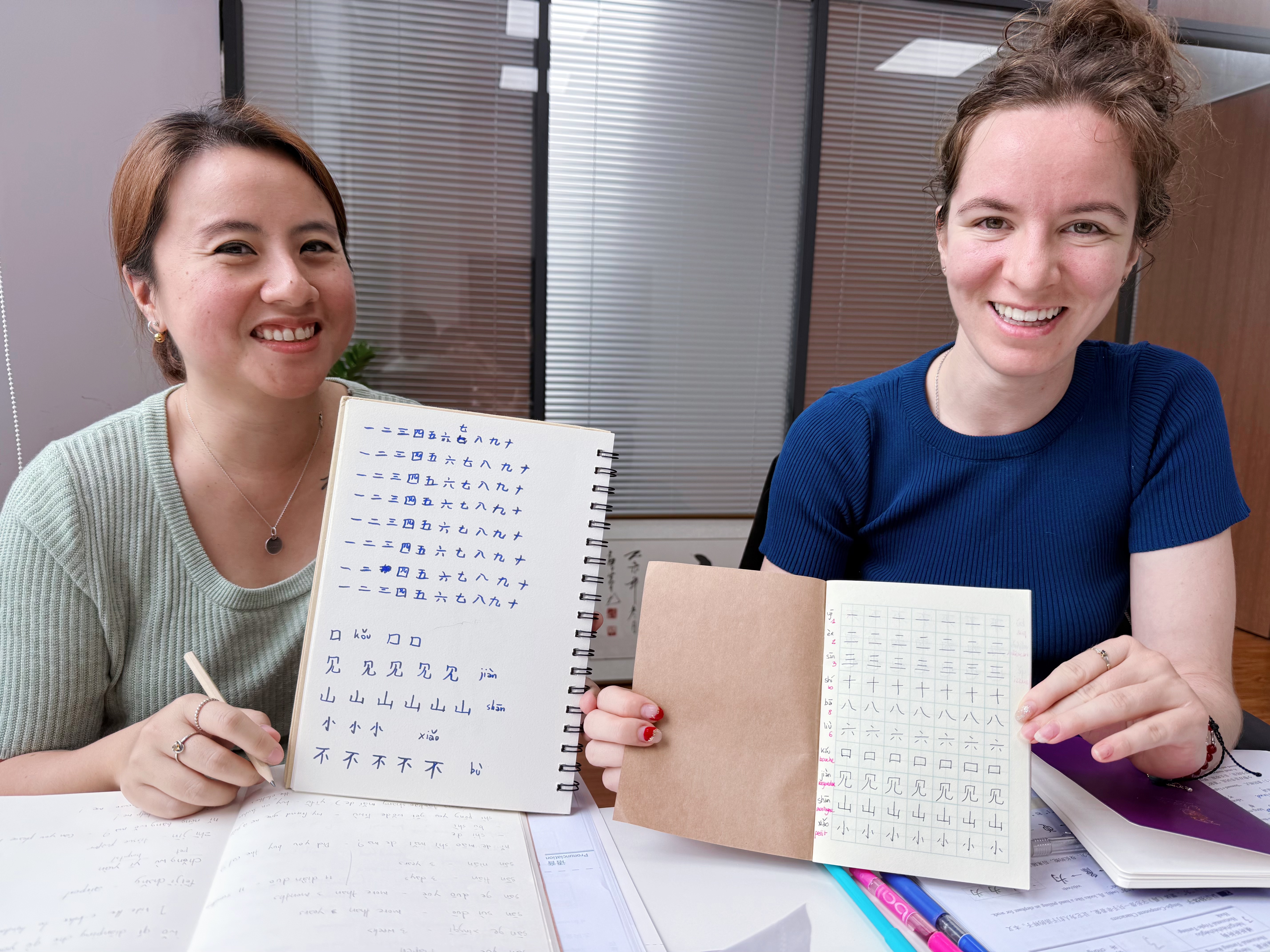 Two women holding up Chinese character repetition booklets, smiling and looking at the camera